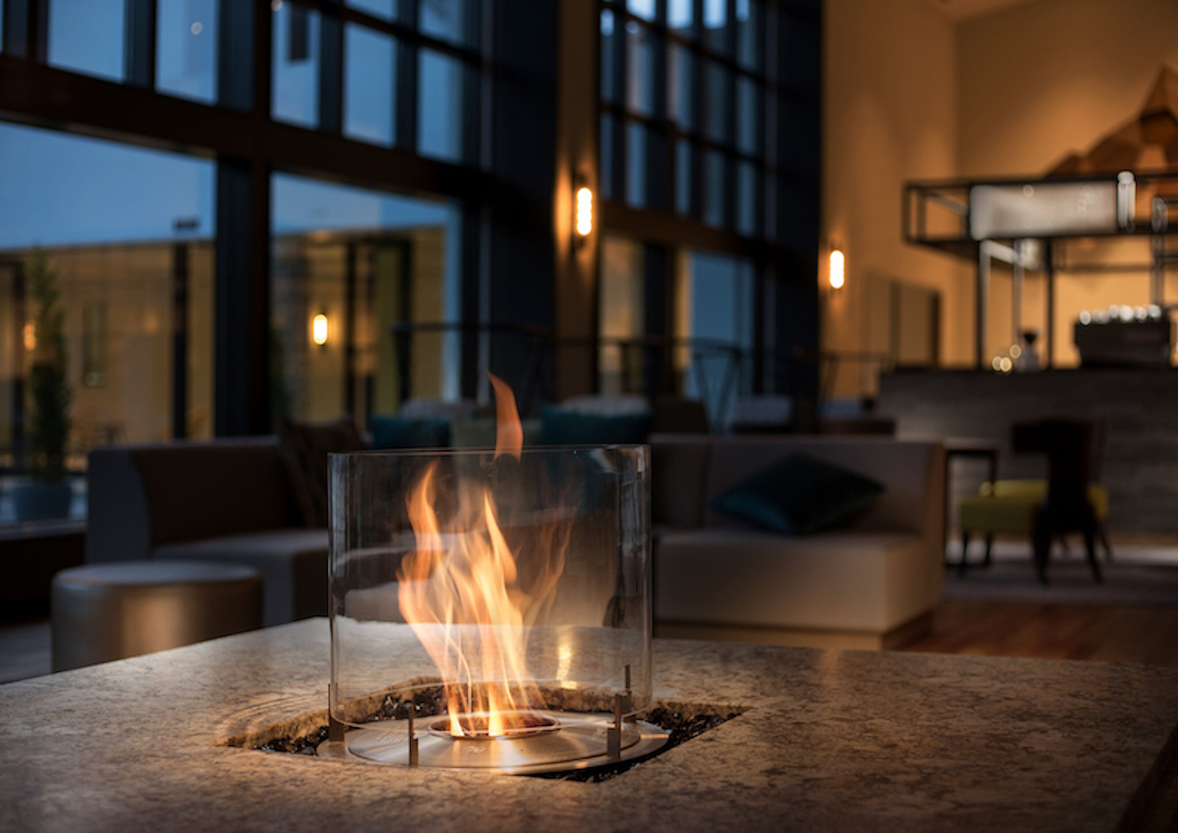A glass table with a fire burning on it, set in a modern interior with a sofa and a dining area in the background.