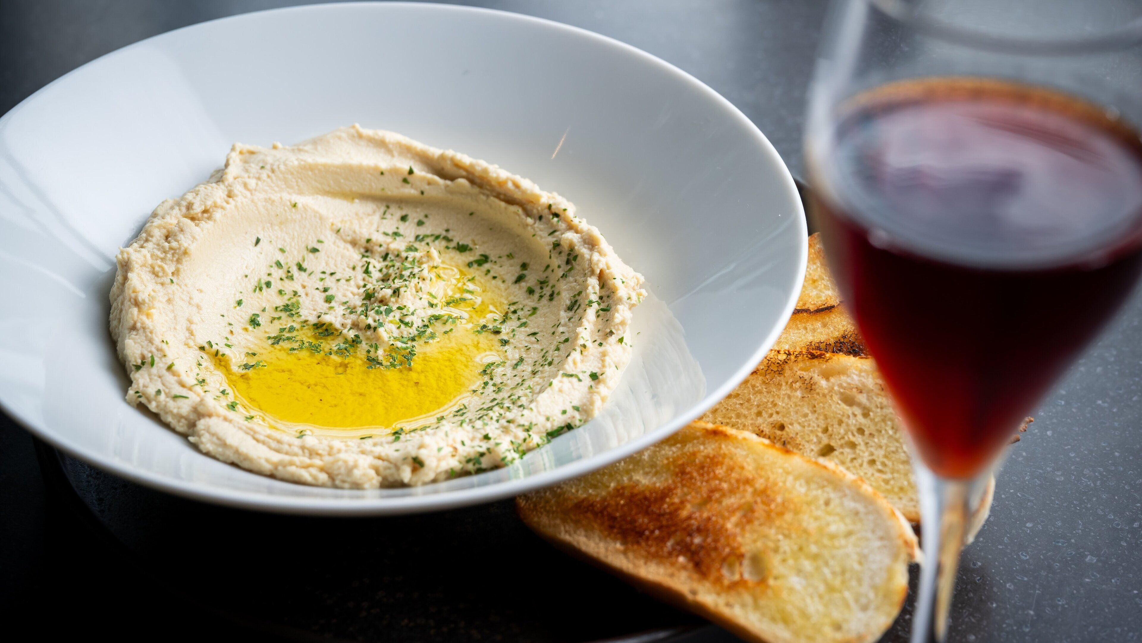 A bowl of hummus with olive oil and herbs, accompanied by toasted bread and a glass of red wine.
