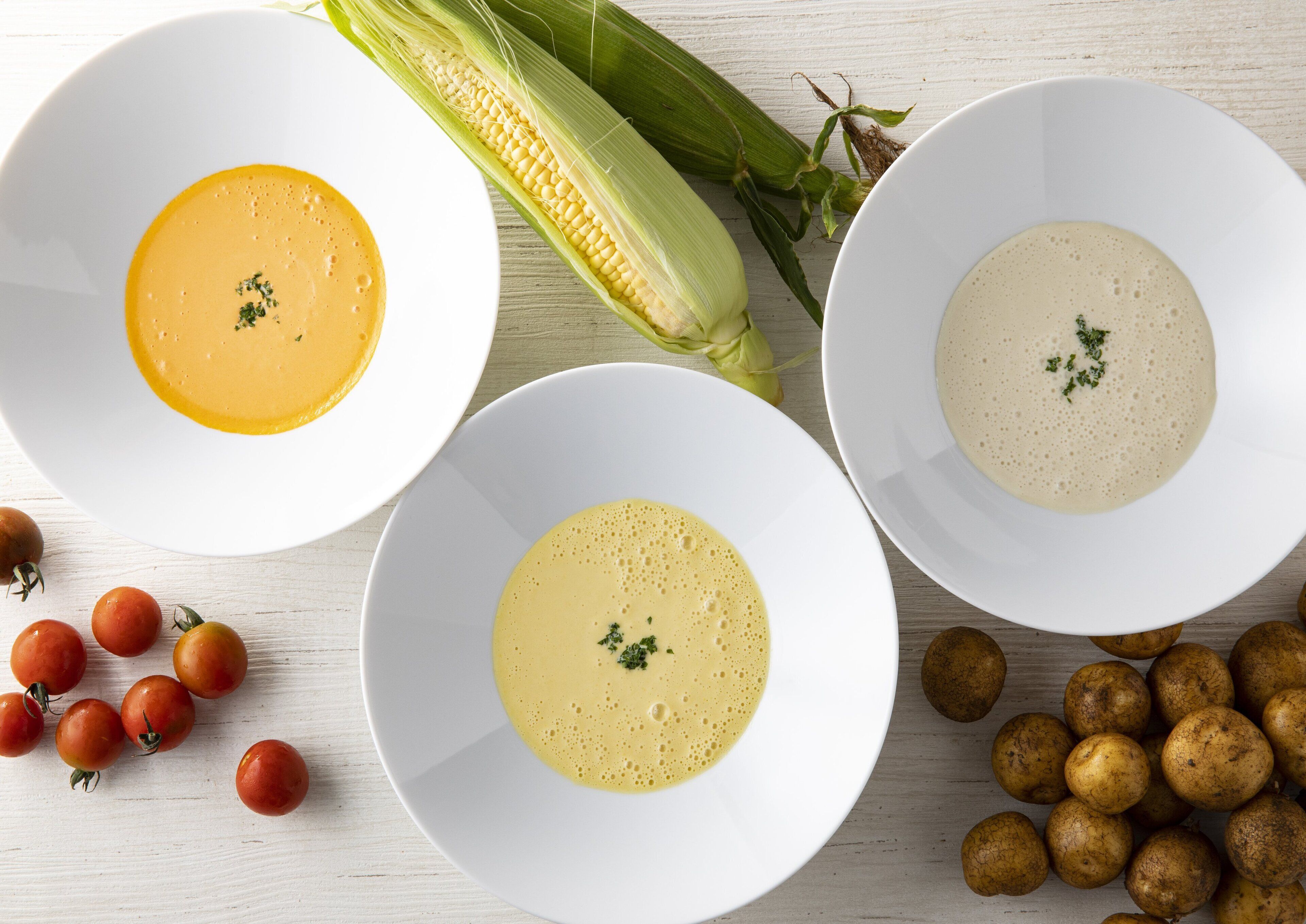 Tres tazones blancos de sopa, adornados con una mazorca de maíz, un manojo de tomates cherry y patatas sobre una superficie de madera.