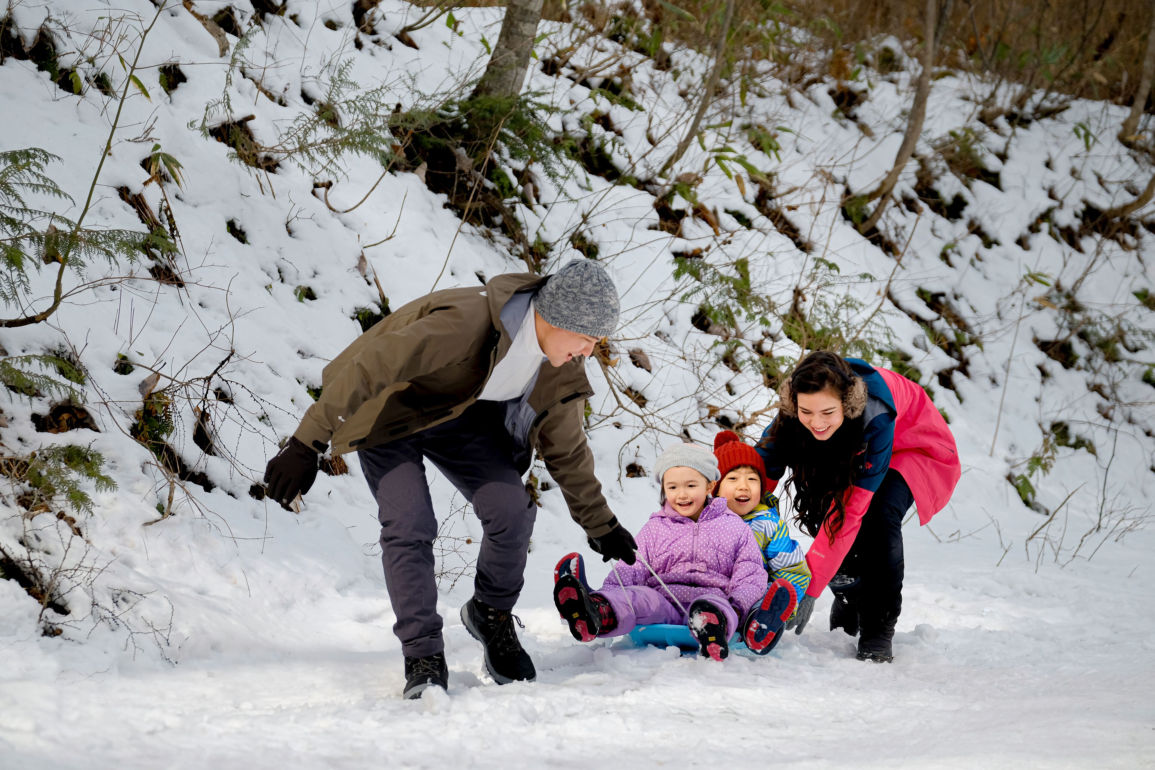 Une scène hivernale en plein air, enneigée, où l’on voit une personne et deux enfants faire de la luge.