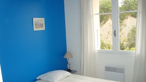 A room with two beds, a window with curtains, a radiator, and a small table with a lamp. - Detached 3-star village house overlooking the Durance river. (Thèze)