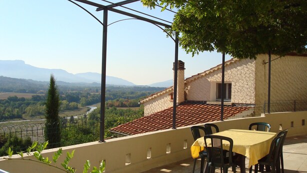 A terrace with a table and chairs, a view of the countryside, and a building with a tiled roof. - Detached 3-star village house overlooking the Durance river. (Thèze)
