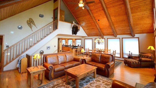 A spacious living area with a high ceiling, wooden beams, a staircase, leather sofas, a dining table, and a mounted moose head. - Morning Glory - 3 bedroom home - quiet wooded location (Estes Park)