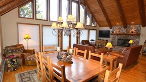 A spacious living room with a wooden dining table, chairs, a fireplace, and a stone wall. - Morning Glory - 3 bedroom home - quiet wooded location (Estes Park)