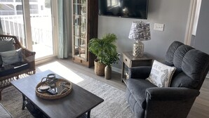 A living room with a sofa, a coffee table, a television, a bookshelf, and a window with curtains. - Gulf Shores Plantation Resort by ALBVR (Gulf Shores)