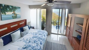 Une chambre à coucher avec un lit, un ventilateur de plafond, un tableau, une armoire et une vue sur l’extérieur.