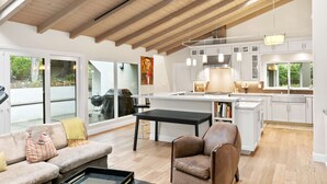 Amplia sala de estar con un sofá, una mesa de centro y un sillón marrón. Se observa una cocina con armarios blancos y una isla grande.