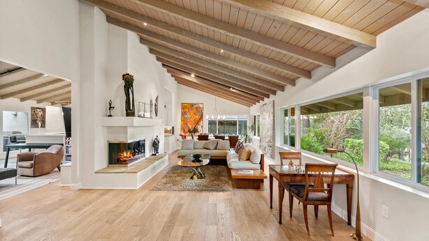 Amplio salón con chimenea, suelo de madera y ventanas grandes que ofrecen vistas al verdor.