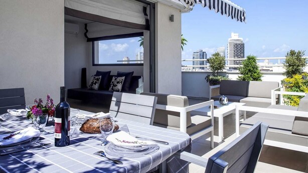 Eine Dachterrasse mit Esstisch für zwei Personen, einer Weinflasche und einem Brot. Gepolsterte Sitzbereiche und Blick auf die Stadtlandschaft.