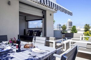 A rooftop terrace with a dining table set for two, a bottle of wine, and a loaf of bread. There are cushioned seating areas and a view of the city skyline. - Duplex Penthouse by Rothschild Pkg TL54 (Tel Aviv)