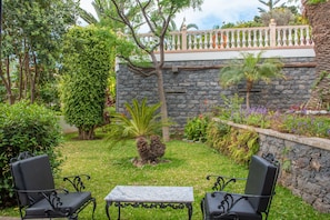 Um jardim com muro de pedra, duas cadeiras de metal preto, uma mesa de mármore e diversas plantas.