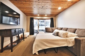 Un salón moderno con techo de madera, un televisor de pantalla plana montado en la pared, un sofá con cojines, una mesita con una lámpara y vistas a través de un ventanal.