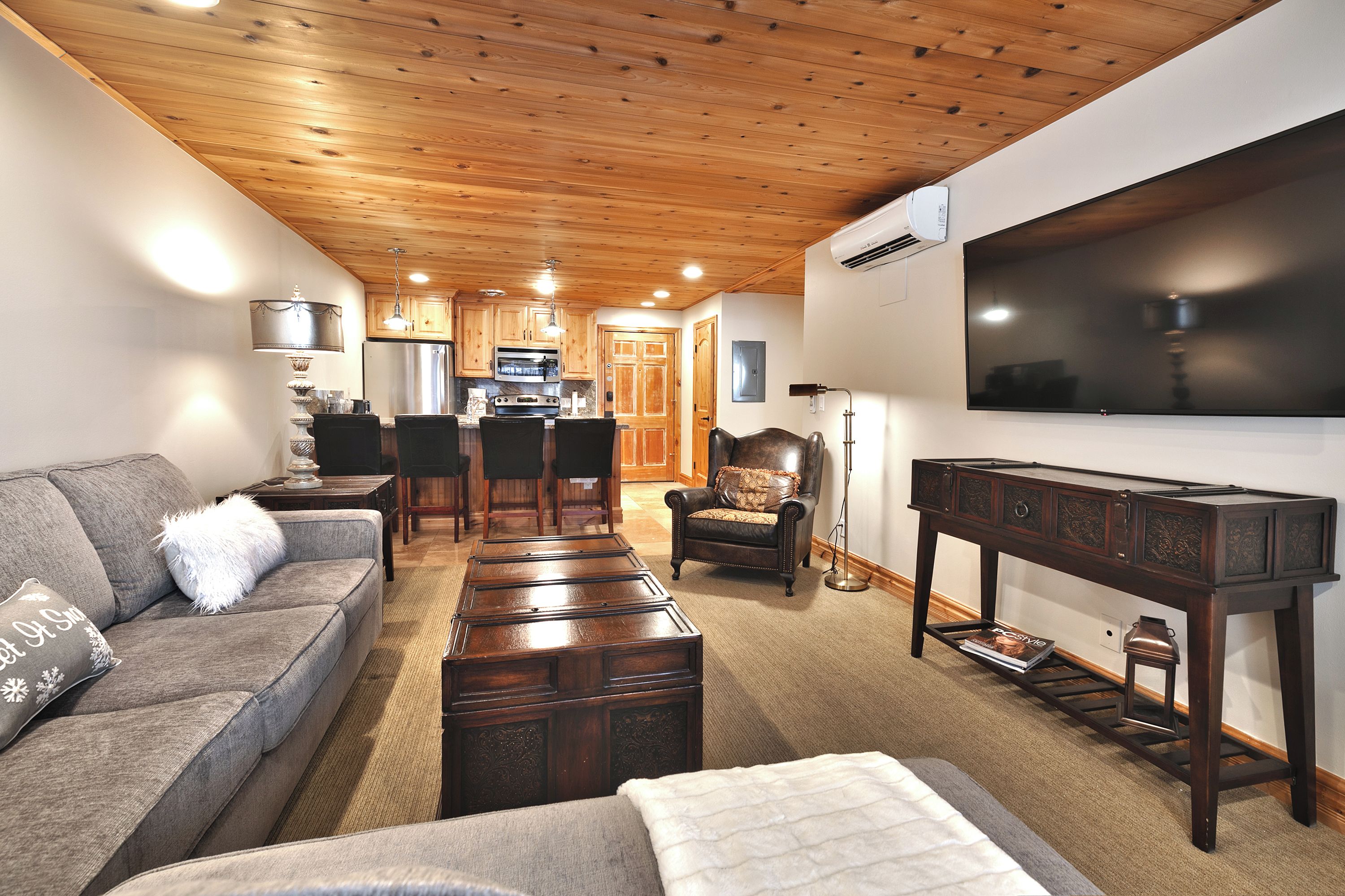 A living room with a wooden ceiling, a sofa, a coffee table, a TV, and a kitchen in the background.