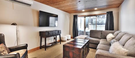 A living room with a wooden ceiling, a flat-screen TV mounted on the wall, a leather armchair, a sofa with fluffy cushions, and a dark wooden coffee table.
