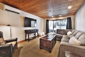 Un salon avec un plafond en bois, un téléviseur à écran plat fixé au mur, un fauteuil en cuir, un canapé avec des coussins moelleux et une table basse en bois foncé.