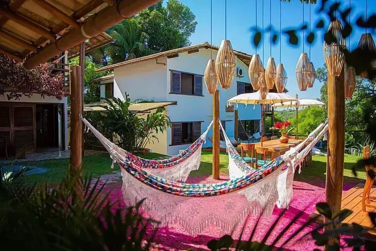 A hammock under a wooden pergola, a dining area with tables and chairs, and a white building with windows in the background.