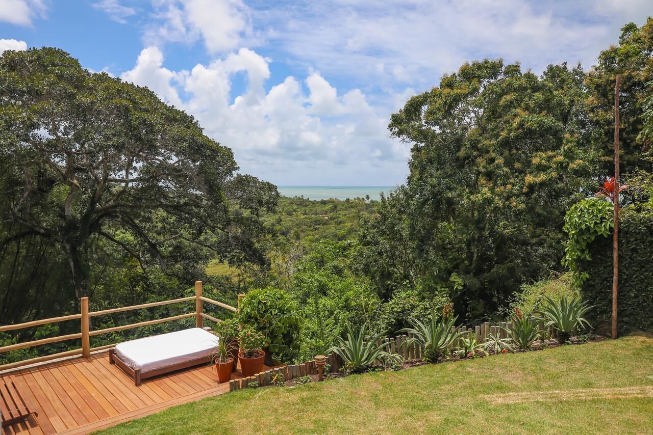 Una terraza de madera con una cama individual, rodeada de abundante vegetación y vistas a una costa distante.