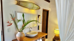 Un baño con tocador de madera, un lavamanos blanco, un espejo, una planta en maceta y un perchero para toallas.