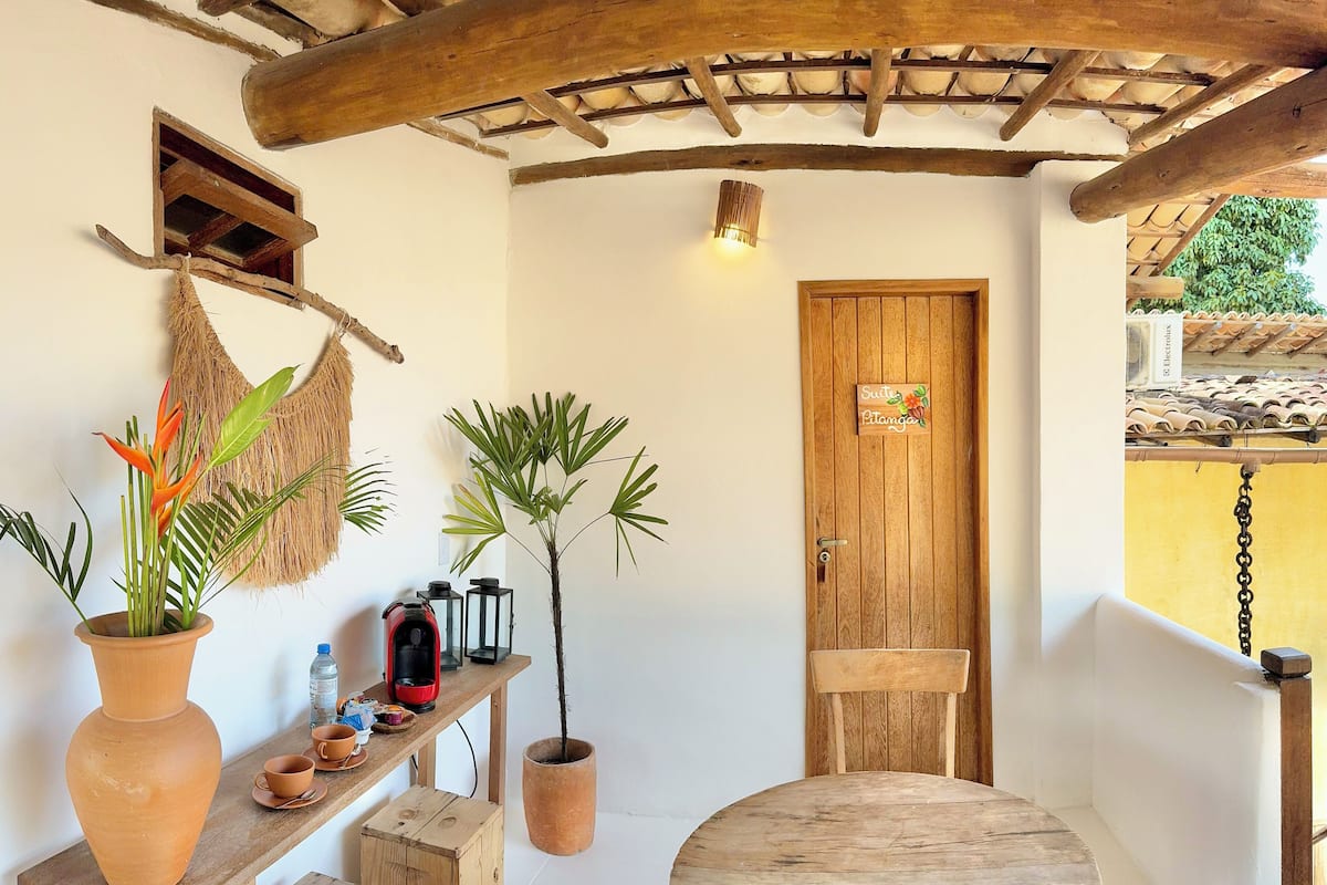 A rustic room with a wooden table, a potted plant, a small stool, a shelf with a coffee maker, and a door with a sign.
