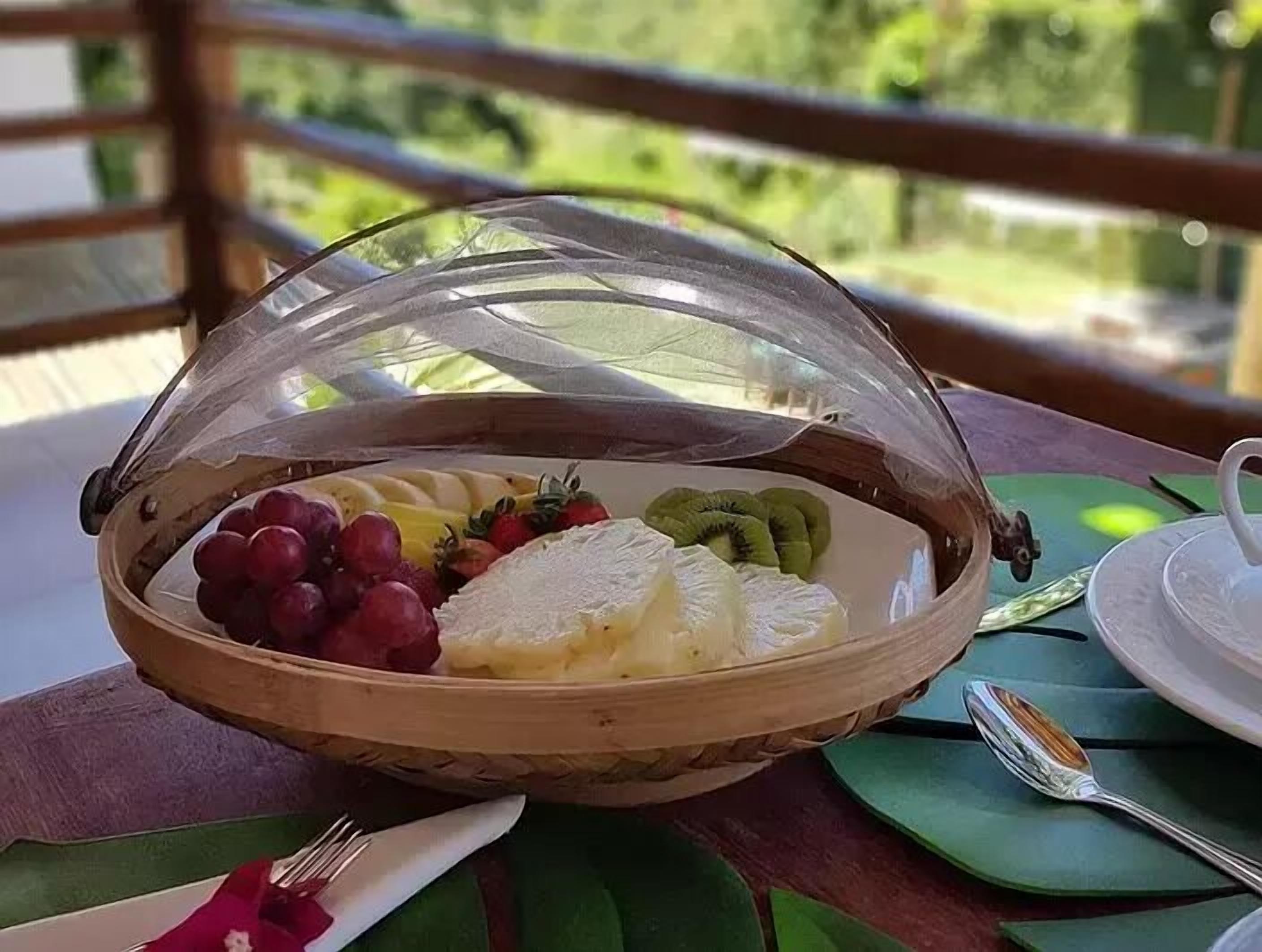 Uma mesa de madeira com uma cesta de frutas coberta por uma cúpula de vidro, um prato branco com fruta fatiada e uma colher.