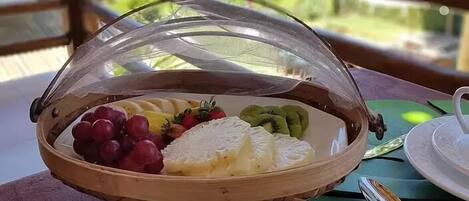 Uma mesa de madeira com uma cesta de frutas coberta por uma cúpula de vidro, um prato branco com fruta fatiada e uma colher.