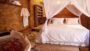 Un dormitorio con una cama grande, cabecera de madera, bañera con pétalos de rosa, estante con toallas y pared de ladrillo.