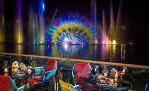 A nighttime view of a water fountain show with colorful lights, a dining area with chairs and tables, and a bar with drinks.