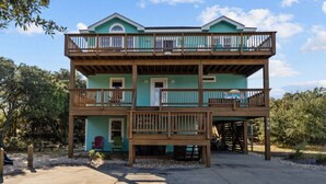 A two-story building with a balcony on the upper level, a covered porch on the ground floor, and a small outdoor seating area. - Family Fun House! Private Pool, Putting Green, Walk to Beach, Hot Tub, Game Room (Corolla)
