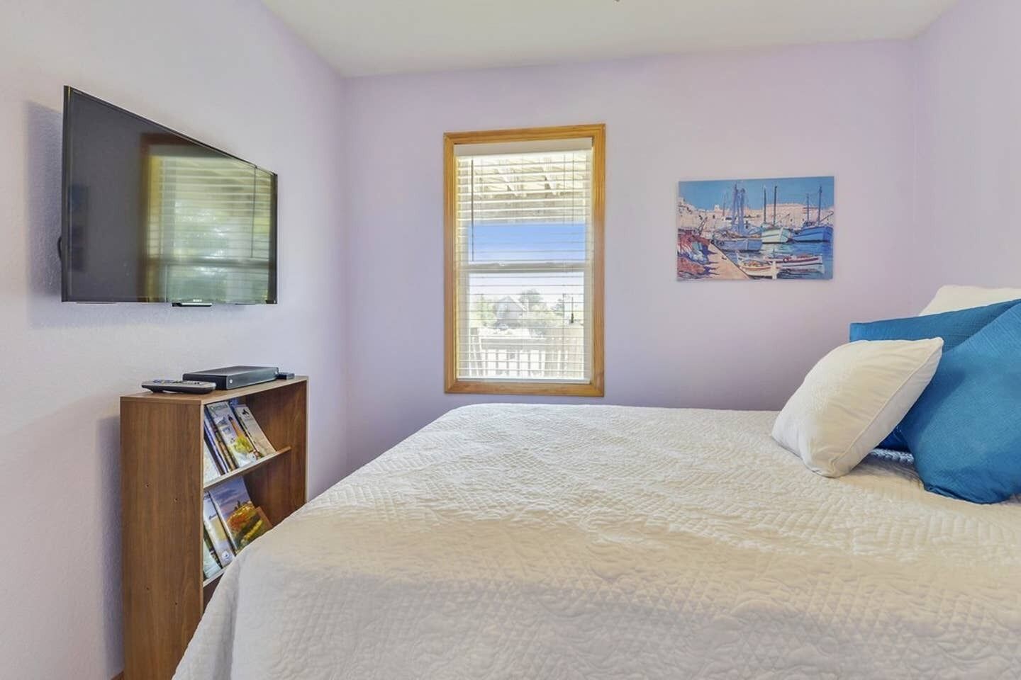 Une chambre à coucher avec un lit, un téléviseur fixé au mur, une fenêtre avec des stores, une bibliothèque et un tableau.