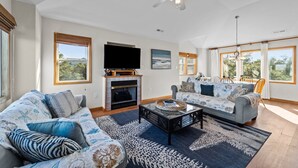 A spacious living room with a fireplace, a ceiling fan, and large windows offering views of the outdoors. - Family Fun House! Private Pool, Putting Green, Walk to Beach, Hot Tub, Game Room (Corolla)