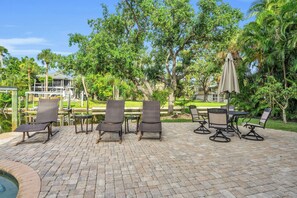An outdoor patio with wicker lounge chairs and a table, surrounded by lush greenery and a pool. - spectacular architectural home steps to the beach w/private heated pool on canal (Fort Myers Beach)