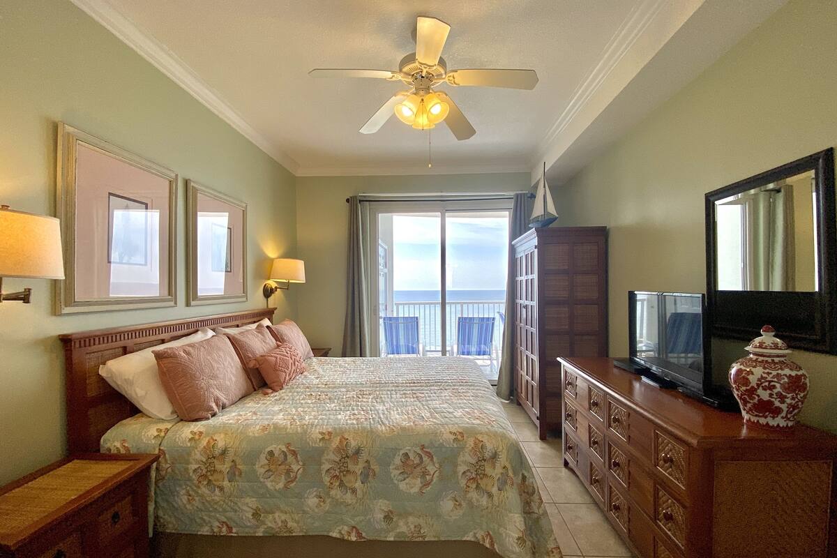 A hotel room with a bed, a ceiling fan, a television, a dresser, and a balcony with a view.