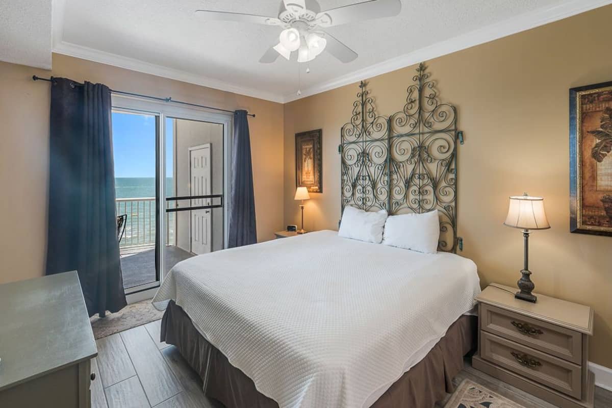 A bedroom with a large bed, a ceiling fan, a nightstand with a lamp, and a view of the ocean through a sliding door.