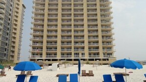 Un hôtel de grande hauteur doté de nombreux balcons, une plage de sable fin avec des chaises longues bleues et des parasols, et une vue dégagée sur l’océan.