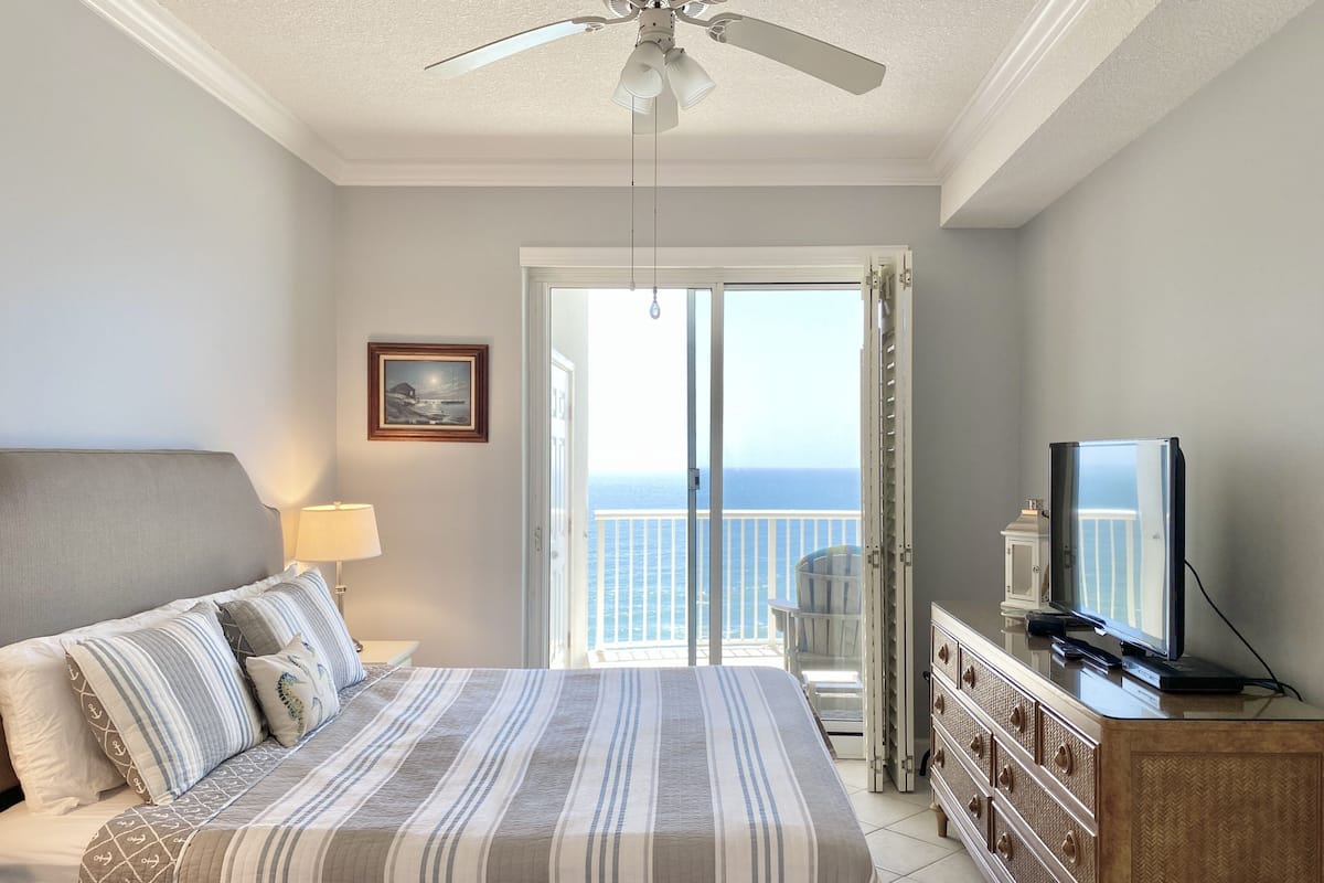 A hotel room with a bed, a television, a dresser, and a balcony with a view of the ocean.