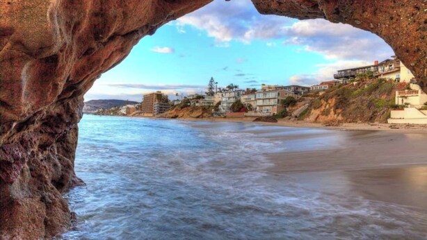 A coastal view through a natural rock arch with a beach and residential buildings in the background. - No Stairs! Walk 2 Ocean, FastWiFi, PoolTable, Magnolia Perch! STR14-0085 (Dana Point)