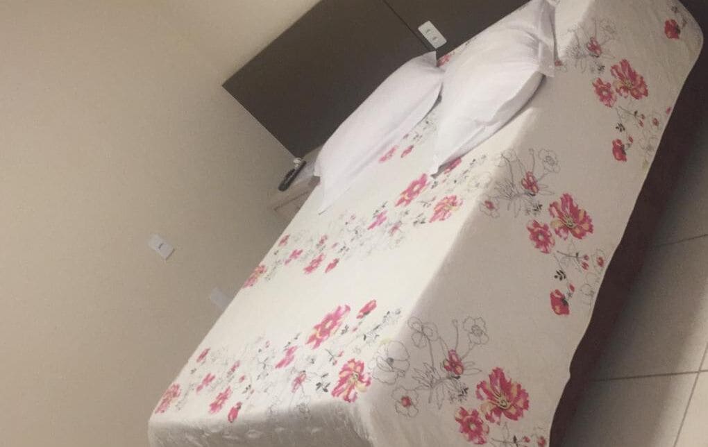 Uma cama com colcha florida e travesseiros brancos num quarto com parede lisa e cabeceira escura.