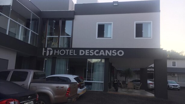 Ein modernes Hotel mit einem Schild, auf dem 'HOTEL DESCANSO' steht. Vor dem Hotel sind mehrere Fahrzeuge geparkt, darunter ein Pickup und eine Limousine.