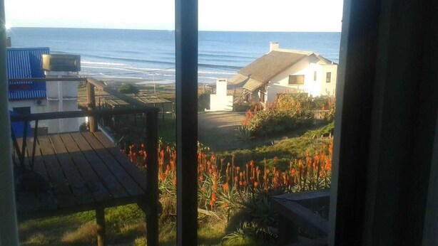 Une vue depuis une fenêtre donnant sur un jardin où l’on aperçoit des fleurs rouges, une terrasse en bois et un bâtiment dont le toit est recouvert de chaume.