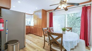 A kitchen with wooden cabinets, a dining table with chairs, a ceiling fan, and a window with red curtains.
