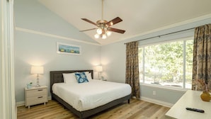 A bedroom with a bed, nightstands, a ceiling fan, a window with curtains, and a view of greenery outside.
