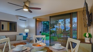 A dining area with a glass table, white chairs, and a bowl of fruit. There is a ceiling fan above and a TV mounted on the wall. - Gorgeous Ground Floor Oceanfront Condo, Just Steps Away From Pool & Beach! (Kihei)