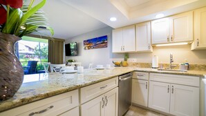 A modern kitchen with white cabinets, granite countertops, and stainless steel appliances. There is a dining area with a table and chairs visible through a glass door. - Gorgeous Ground Floor Oceanfront Condo, Just Steps Away From Pool & Beach! (Kihei)