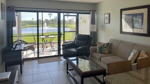 Un salon avec une table en verre, un canapé en cuir, un fauteuil noir et une vue sur un plan d’eau et la verdure, grâce à de grandes portes coulissantes.