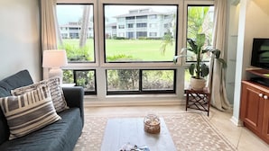 A living room with a sofa, a coffee table, a TV stand, and large windows offering a view of a building and greenery. - CC 1311 Golf Course View Condo - Paradise Vacation Rental in Ocean Village (Fort Pierce)
