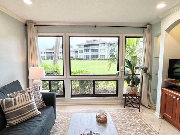 A living room with a sofa, a coffee table, a TV stand, and large windows offering a view of a building and greenery. - CC 1311 Golf Course View Condo - Paradise Vacation Rental in Ocean Village (Fort Pierce)