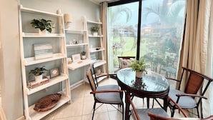 A dining area with a round glass table, surrounded by wooden chairs, and flanked by white shelving units with plants and decorative items. - CC 1311 Golf Course View Condo - Paradise Vacation Rental in Ocean Village (Fort Pierce)