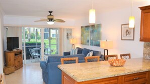 A modern kitchen with a granite countertop, wooden cabinets, and a dining area with a blue sofa and wicker chairs. - OV 617 Pond/Golf Course View Condo-Paradise Vacation Rental in Ocean Village (Fort Pierce)