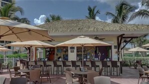 An outdoor café with tables and chairs under large umbrellas, a bar area with a thatched roof, and palm trees in the background. - CP 315 Dune View Condo -Paradise Vacation Rental in Ocean Village (Fort Pierce)