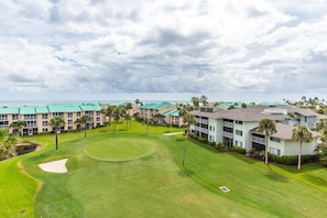 A golf course with a putting green and sand bunkers, surrounded by multi-story buildings with green roofs and palm trees.